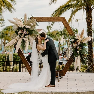 Captured in a tender moment, the bride and groom share a passionate kiss behind a magnificent wooden altar beautifully adorned with bouquets of vibrant flowers. The scene is set against a breathtaking backdrop of a flower-filled oasis, with lush palm trees and glistening water adding to the enchanting ambiance. Created by Simply Enchanted Events, the floral arrangements exude elegance and natural charm, harmonizing perfectly with the surroundings. Love and nature intertwine in this mesmerizing moment at All Inclusive Weddings Orlando.