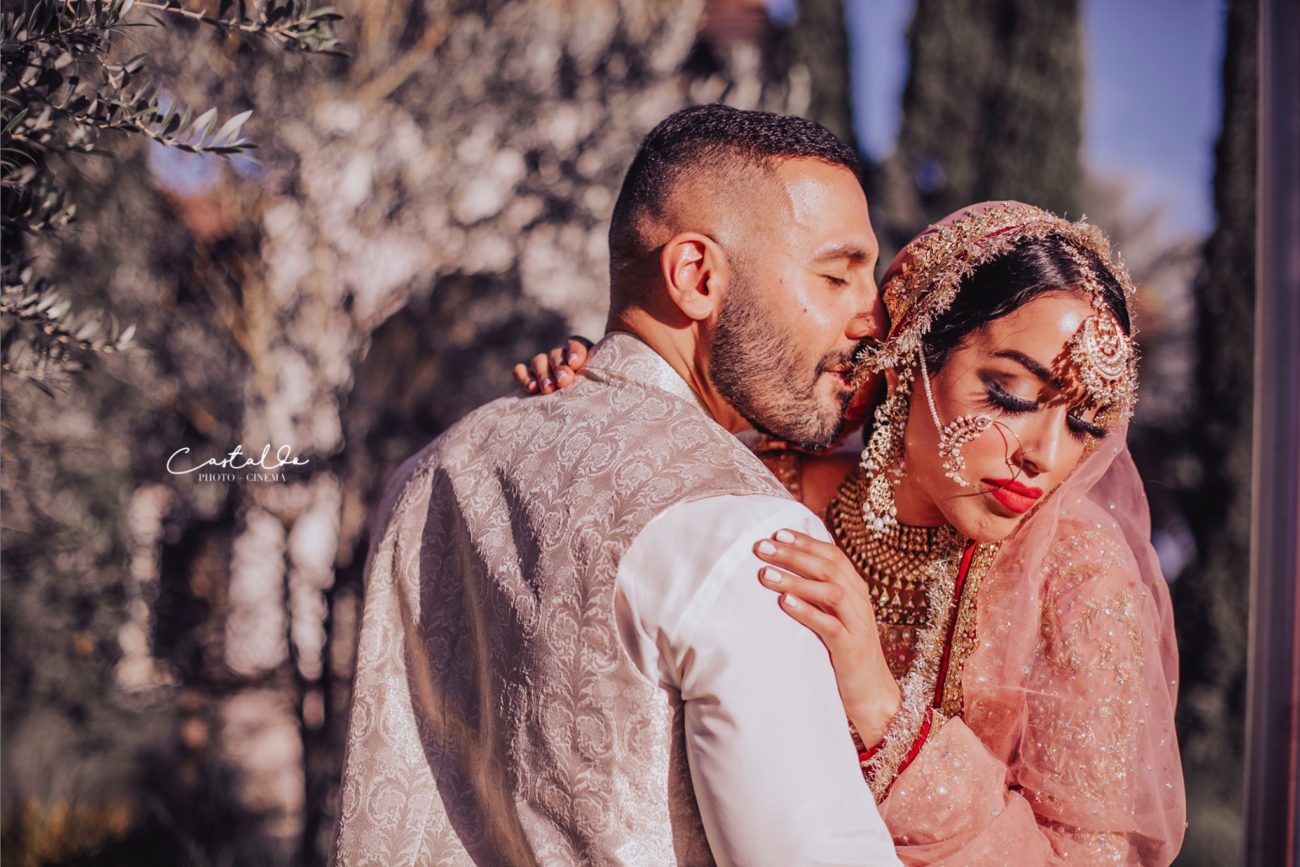 In this captivating photograph by Castaldo Studio, an Indian couple dressed in traditional attire shares an intimate moment. The groom lovingly whispers into his bride's ear, their faces illuminated with joy and affection. The intricate details of their attire and the rich cultural elements add a touch of elegance to the scene. Castaldo Studio expertly captures the tenderness and romance between the couple, creating a truly mesmerizing image. The photo beautifully encapsulates the beauty of their love and the richness of their cultural heritage