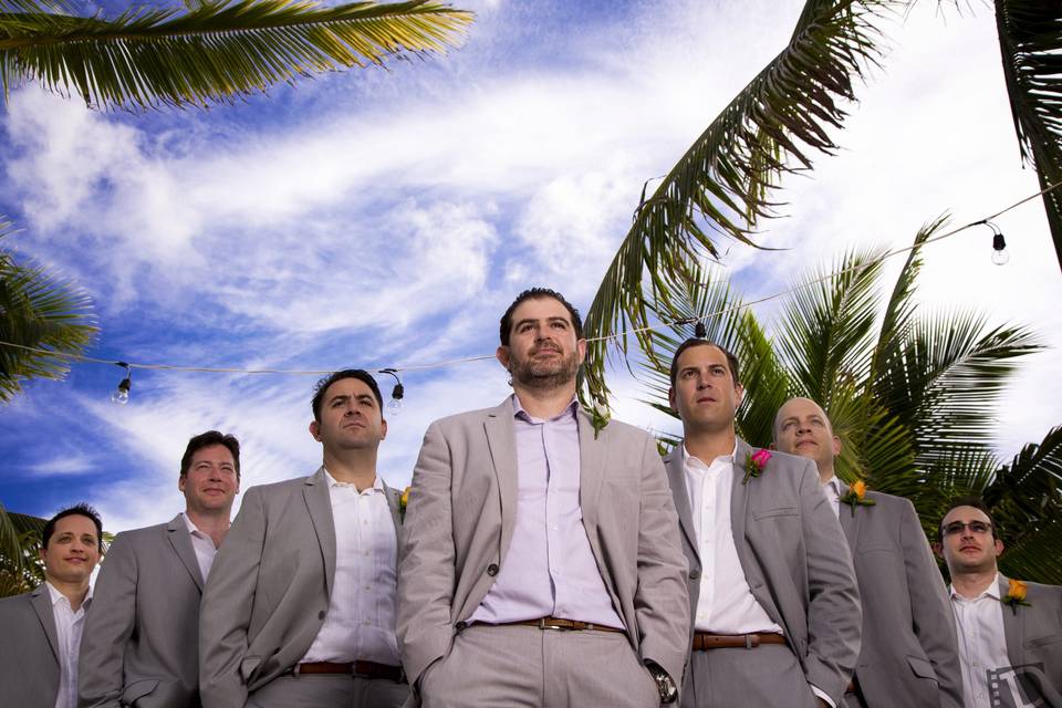 An energetic and playful photo taken by DeMarco Films, featuring the groom confidently leading his groomsmen with a backdrop of swaying palm trees. The vibrant colors and lively atmosphere reflect the excitement and camaraderie of the wedding party