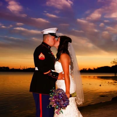 A touching scene on the beach as a military husband kisses his bride, holding a stunning bouquet of purple flowers created by The Flower Studio.