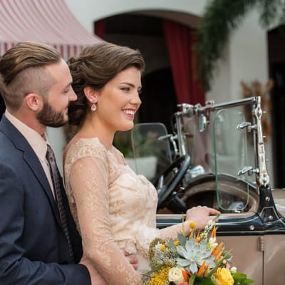 A stylish couple getting into a vintage car, the bride holding a beautiful flower arrangement from The Flower Studio.