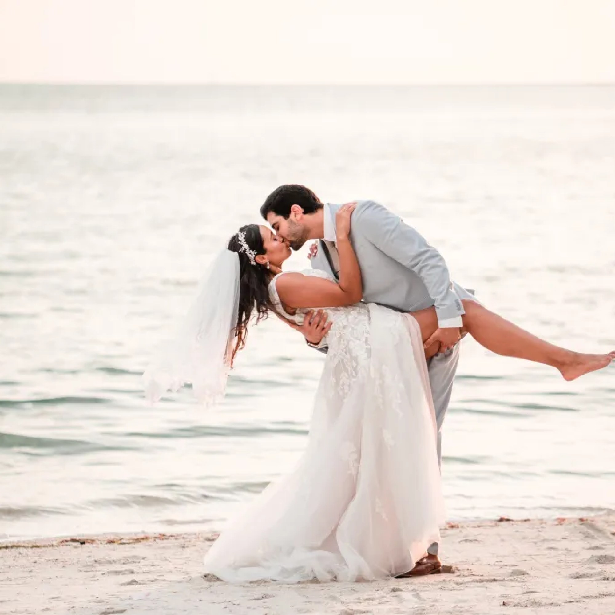 Romantic moment captured at All Inclusive Weddings Orlando, as the groom holds the bride's leg and dips her for a passionate kiss on the beach after their wedding - by Jerzy Nieves Photography