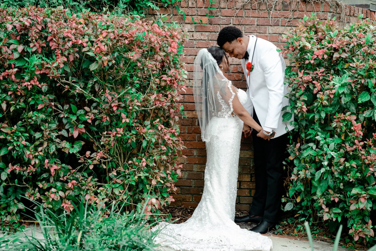 Bride and groom sharing an intimate moment, touching foreheads behind a charming brick wall with a beautiful garden background at All Inclusive Weddings Orlando