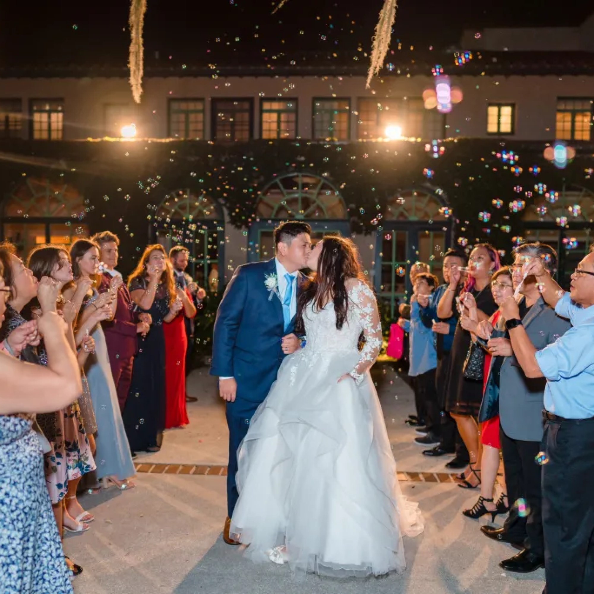 Jerzy Nieves Photography - Newly married couple sharing a romantic kiss while surrounded by their joyful friends and family, who shower them with bubbles, creating a magical and enchanting moment at All Inclusive Weddings Orlando