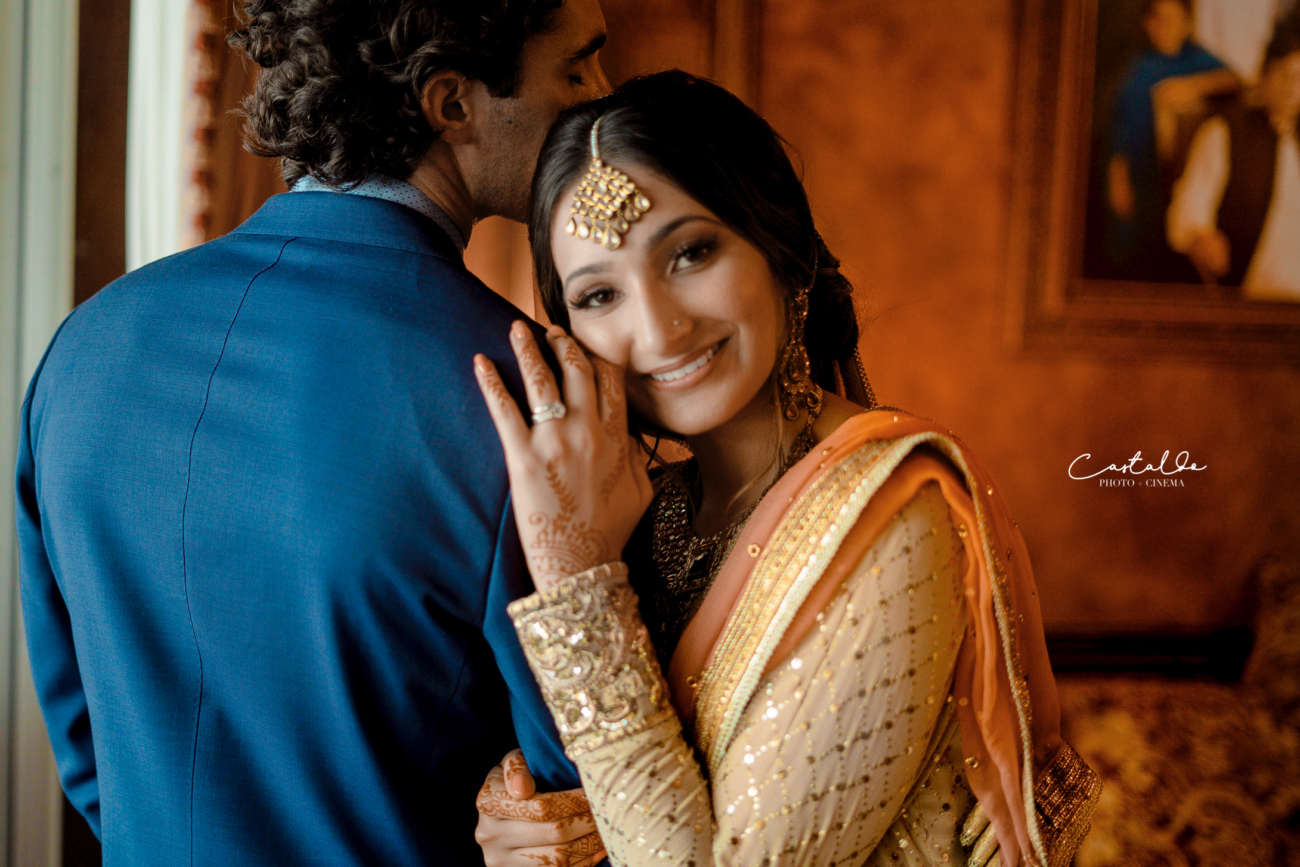 A captivating photograph by Castaldo Studio, showcasing a beaming bride adorned in exquisite traditional Indian wedding attire, embracing her husband. The bride's vibrant and ornate ensemble, along with her radiant smile, beautifully capture the essence of joy and cultural celebration. This picture exudes the rich traditions and the blissful union of the couple on their special day.