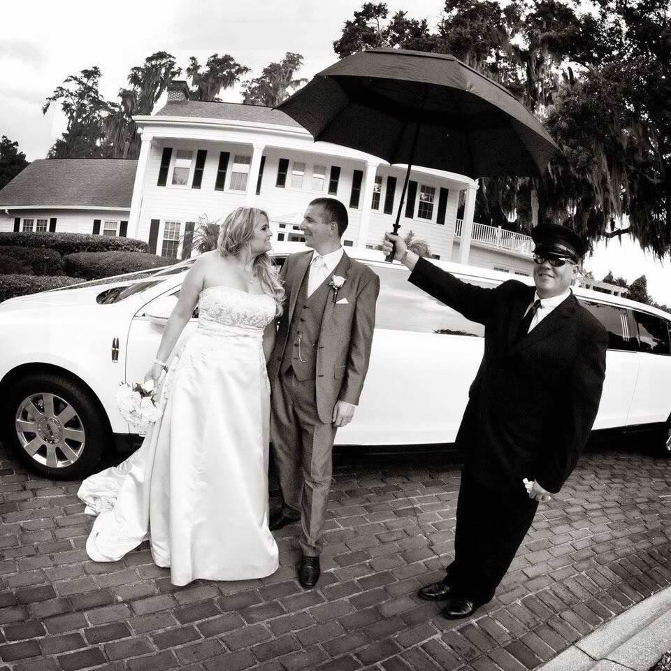 Classic Elegance: Choffer holding umbrella for bride and groom in front of White Stretch Lincoln Limo