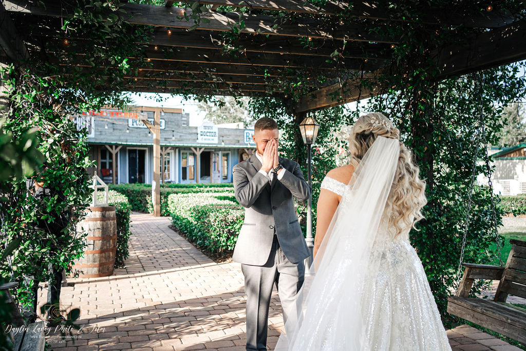 Captured in a tender moment, the bride and groom share a passionate kiss behind a magnificent wooden altar beautifully adorned with bouquets of vibrant flowers. The scene is set against a breathtaking backdrop of a flower-filled oasis, with lush palm trees and glistening water adding to the enchanting ambiance. Created by Simply Enchanted Events, the floral arrangements exude elegance and natural charm, harmonizing perfectly with the surroundings. Love and nature intertwine in this mesmerizing moment at All Inclusive Weddings Orlando.